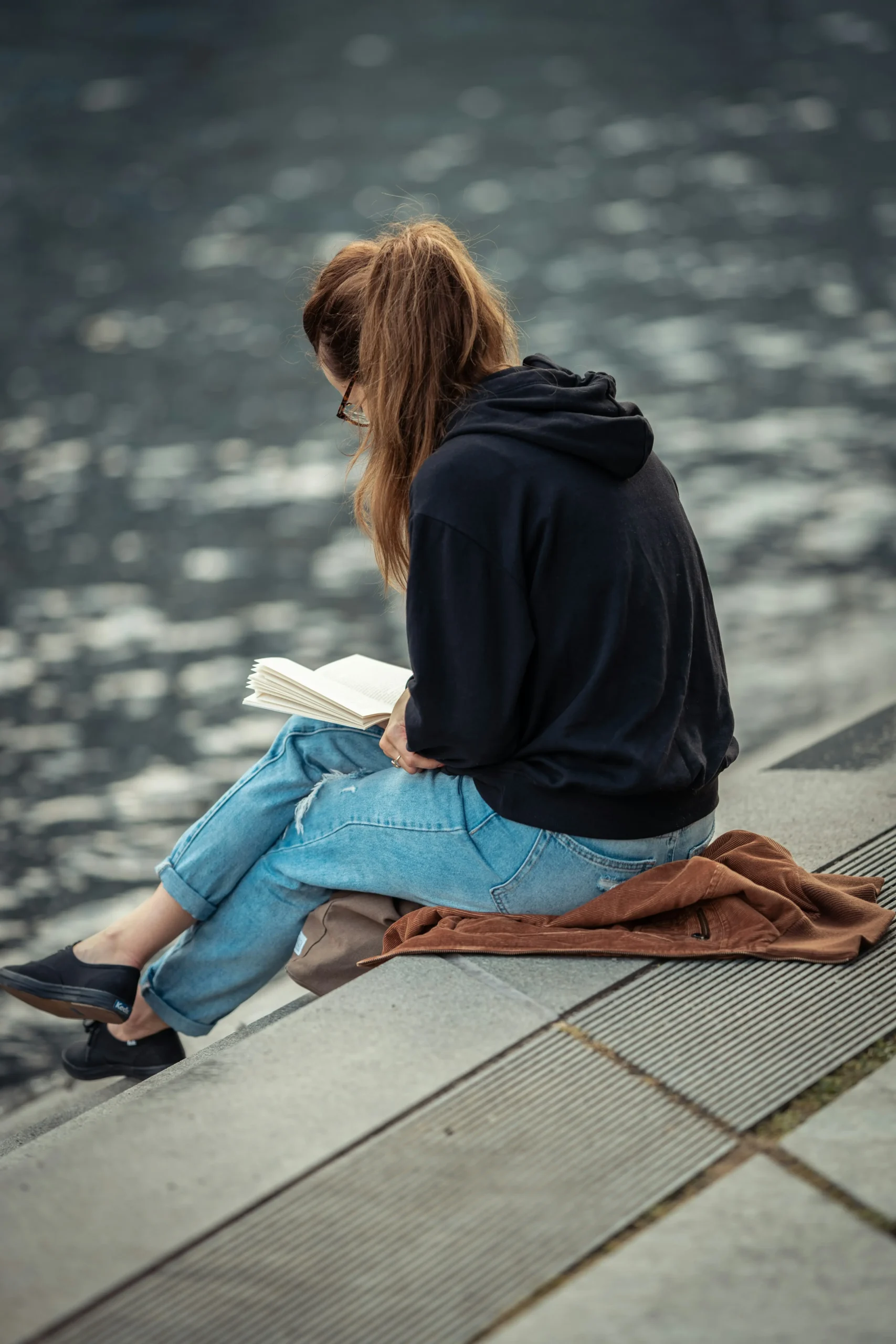 frau-wasser-sitzen-lesen-buch-pulli-hoodie-brille-von-hinten